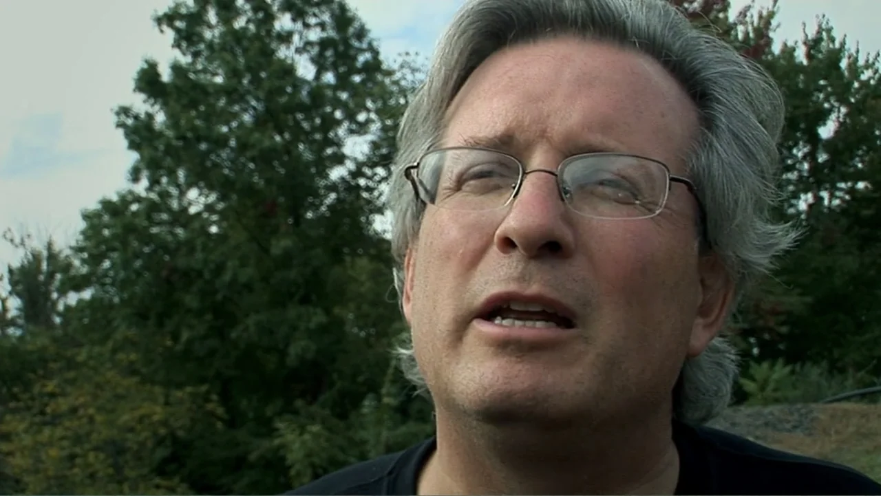 The Cheshire Murders (2013) movie still: closeup — A man is positioned outdoors against a backdrop of blurred green deciduous trees. The man is…