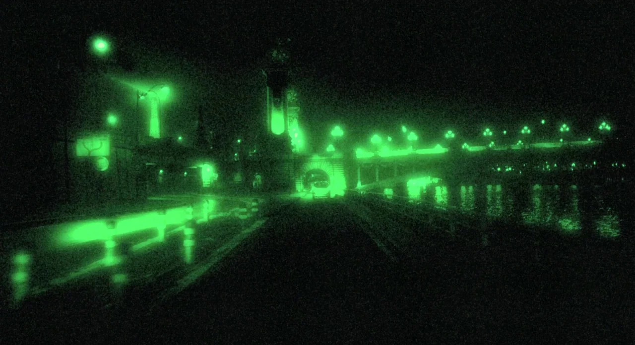 Holy Motors (2012) movie still: establishing — A dark night view of a bridge crossing a river captured on night vision. Multiple streetlights cast…
