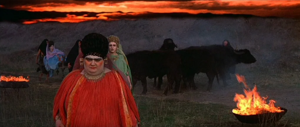 Fellini – Satyricon (1969) movie still: fullbody — A group of people walk through a dark, desolate field at dusk. Large iron braziers containing…