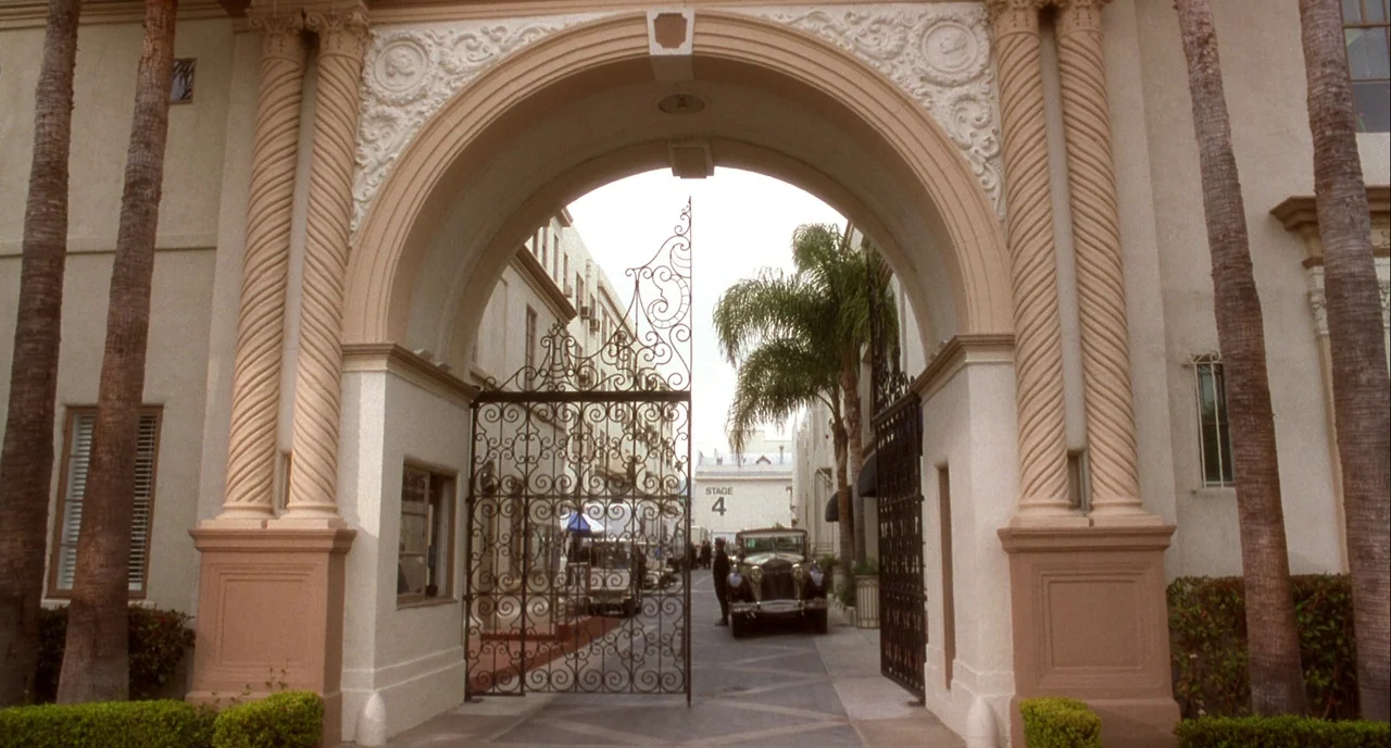 Mulholland Drive (2001) movie still: wide — A formal stone arched entrance with decorative columns leads into a film studio lot. An ornate black…