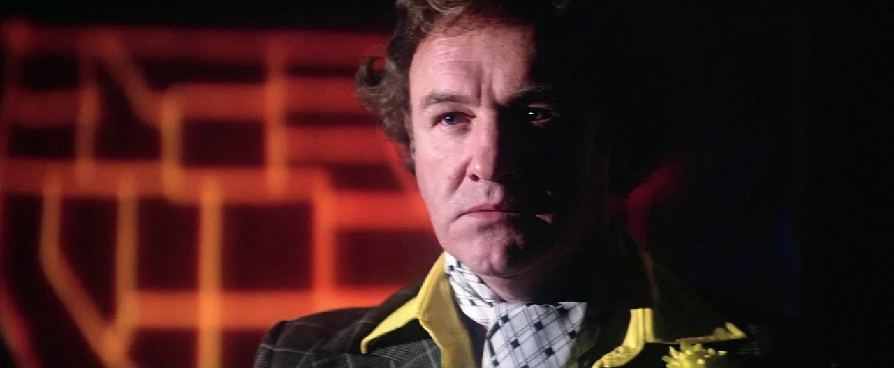 Superman (1978) movie still: closeup — A dimly lit environment features a blurred red grid pattern glowing in the background. A man wearing a plaid…