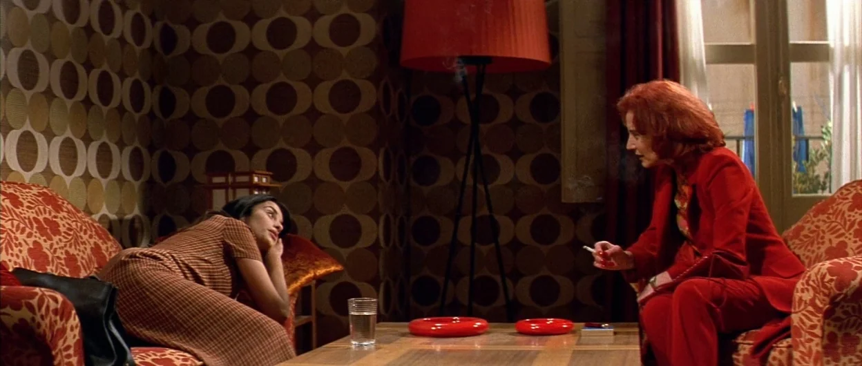All About My Mother (1999) movie still: medium — A living room features 1970s geometric wallpaper and bold floral-patterned upholstery on armchairs.…