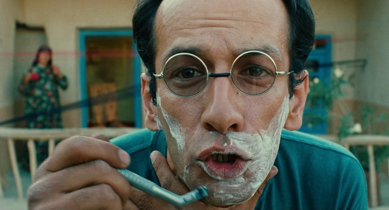 The Wind Will Carry Us (1999) movie still: closeup — A man with shaving cream on his jaw looks directly into the camera lens. In the background a…
