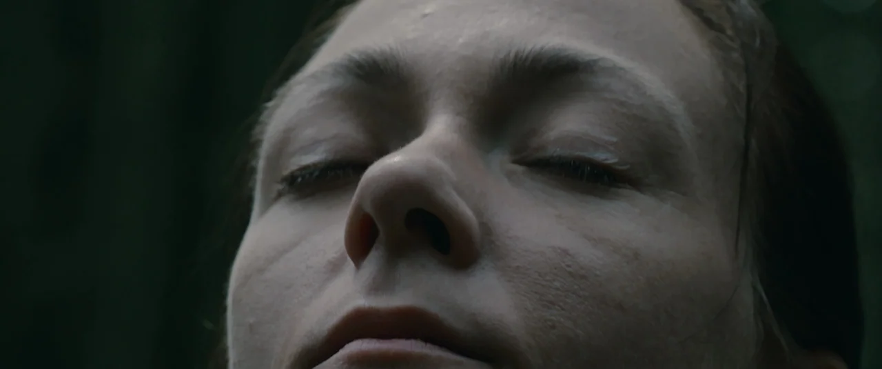 Hagazussa (2017) movie still: closeup — The face of a woman is centered in the frame against an out-of-focus background. She lies with her eyes…