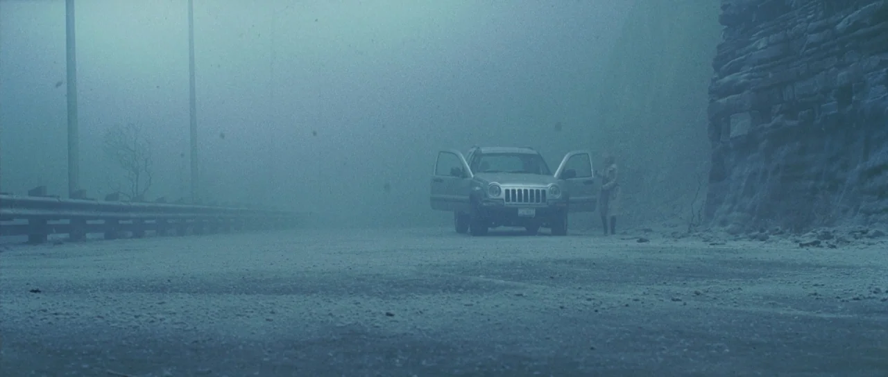 Silent Hill movie still: wide — A desolate road stretches into dense fog alongside a jagged cliff face. A single car sits parked with both front…