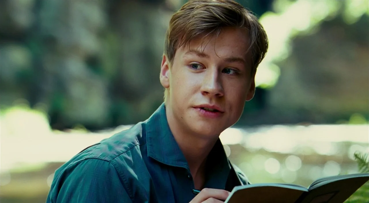 The Reader (2008) movie still: medium — A lush, green outdoor environment with blurred foliage forming the background. A young man sits in the…