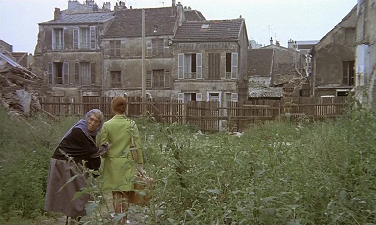 Nuits Rouges (1974) movie still: fullbody — A desolate urban lot overgrown with weeds stands before a row of weathered buildings. Two women walk…