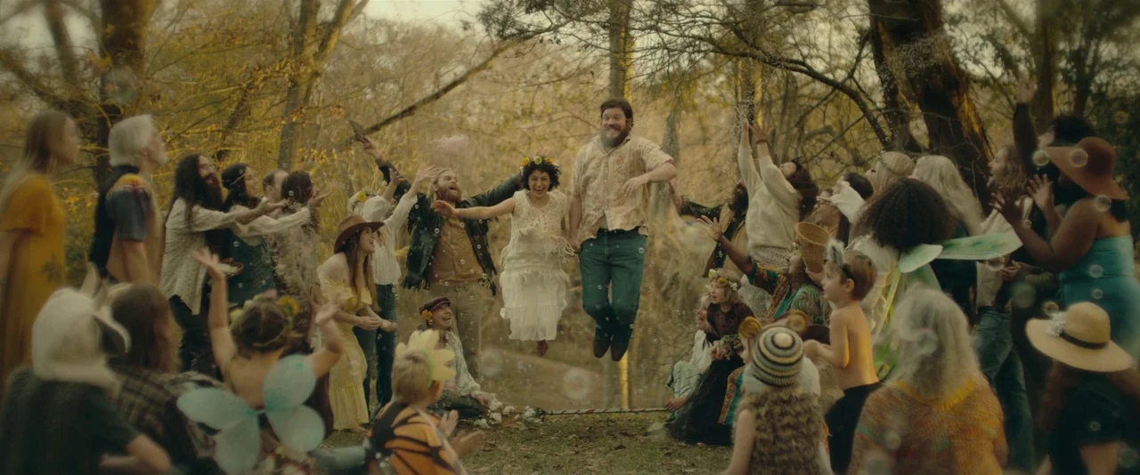 Blaze (2018) movie still: wide — A large group of people gathered in a wooded area during golden hour. A couple jumps in the air while others watch…
