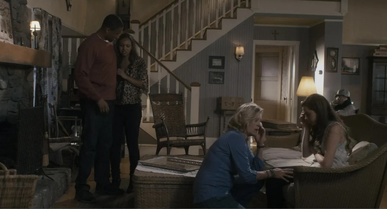 Gates of Darkness (2019) movie still: medium — A living room features a stone fireplace, a wooden staircase, and wicker furniture. Four people stand…