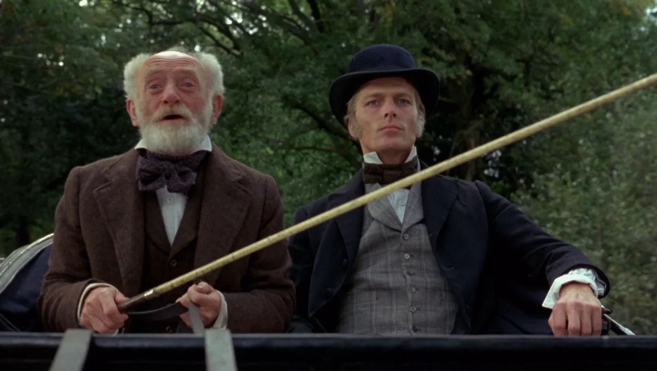 Mountains of the Moon (1990) movie still: medium — Two men sit side by side in a horse-drawn carriage outdoors near dense foliage. The older man…