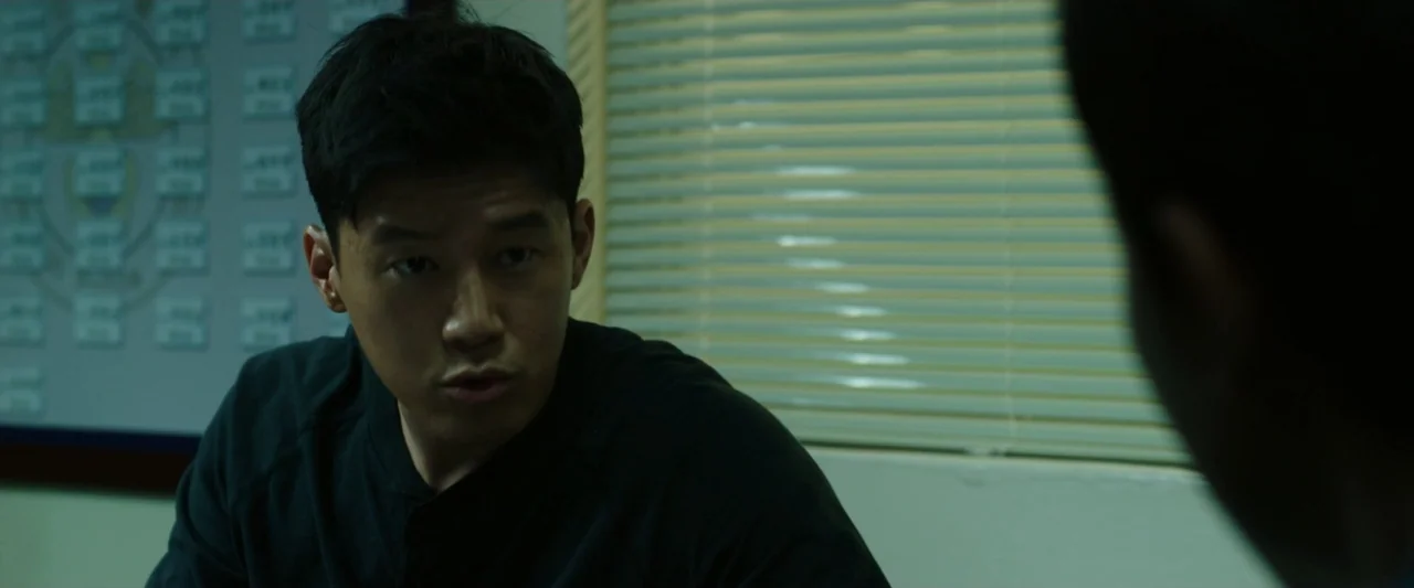 The Gangster, the Cop, the Devil (2019) movie still: medium — An interior office space with horizontal blinds on the window and a wall chart. A…