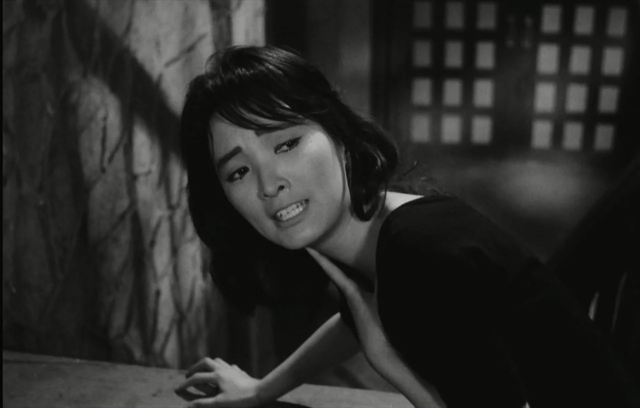 The Housemaid (1960) movie still: closeup — A dark indoor setting features a textured wall and a grid-patterned window or door. A young woman with…
