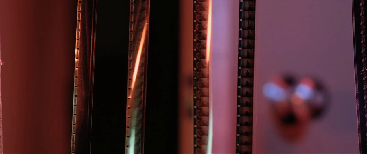 The Editor (2014) movie still: closeup — Vertical strips of developed 35mm film hang in a dark processing room. The film reels catch light, showing…