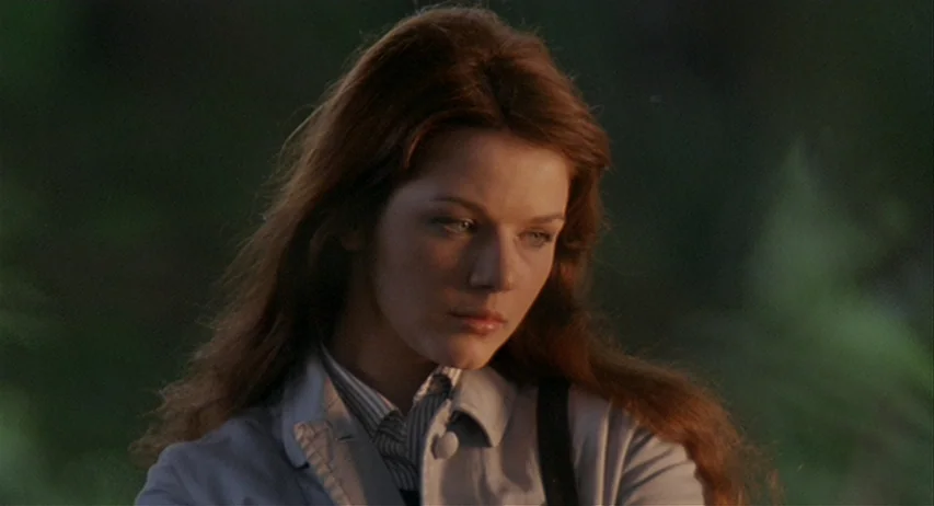 The Fifth Cord (1971) movie still: closeup — An outdoor setting with blurred green foliage visible in the background. A woman with long auburn hair…