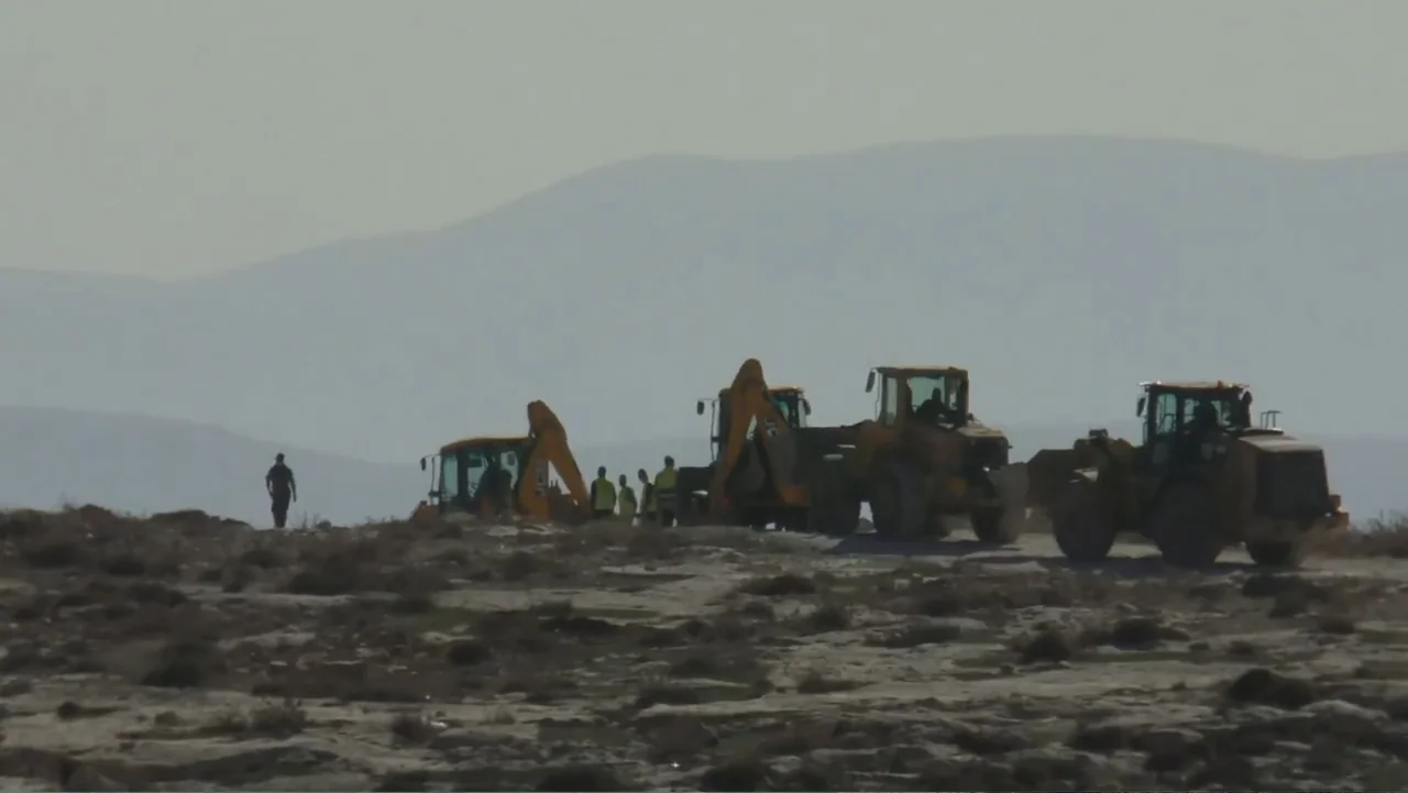 No Other Land (2024) movie still: wide — A group of workers and yellow heavy machinery occupy a rocky hillside clearing. The subjects stand near…