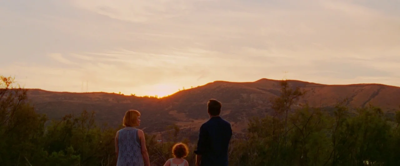 Moon Garden (2022) movie still: wide — A man, woman, and child stand looking out over rolling hills at sunset. The sky is filled with light clouds…