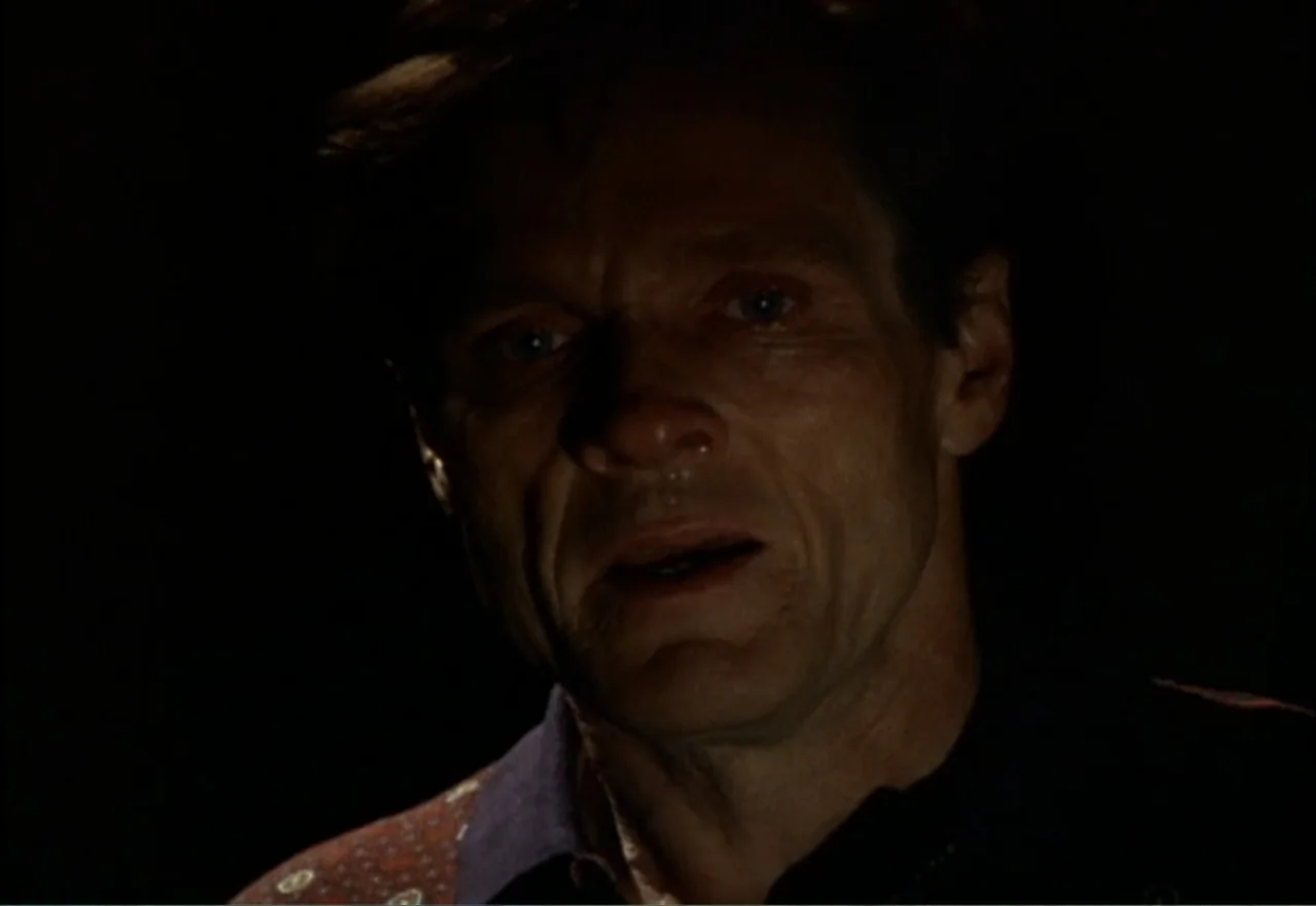 Ambushed (1998) movie still: closeup — A dark, nondescript interior space with the subject illuminated against a black background. A middle-aged man…