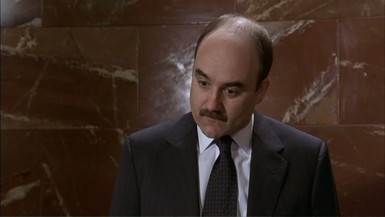 Two Weeks Notice (2002) movie still: medium — A man stands in front of a red polished marble wall in an indoor space. He is wearing a formal suit…