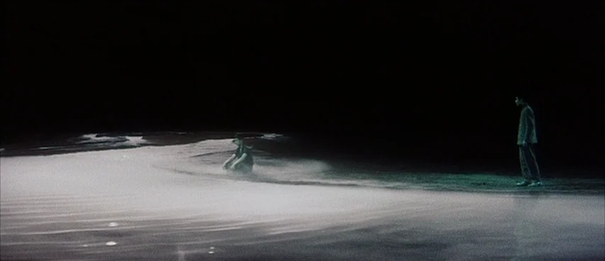 Jigoku (1960) movie still: wide — A dark beach at night with sand illuminated by a concentrated light source. One person is sitting on the sand…