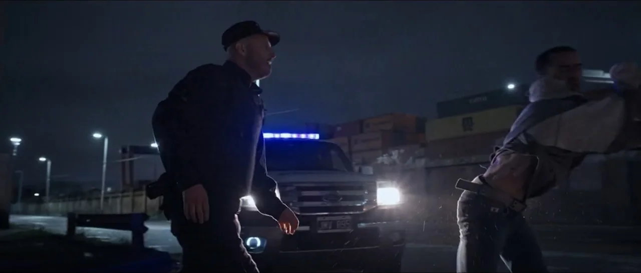 Gunman (2025) movie still: medium — A night scene in a paved lot featuring shipping containers and parked vehicles. One man walks past the front of…