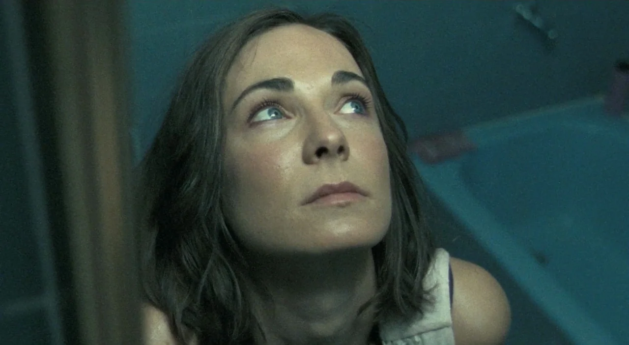 Patagonia (2010) movie still: closeup — A dark, teal-toned space containing a bathtub and tiled walls. A woman looks upwards toward an unseen light…