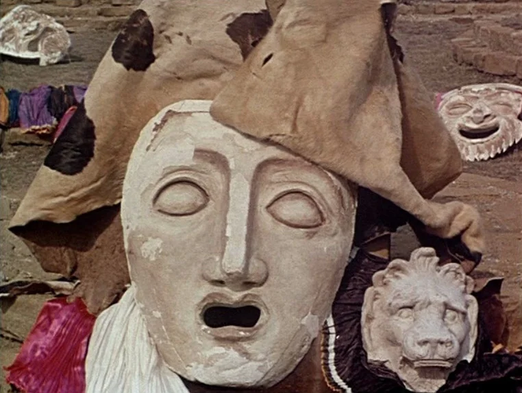Ashik Kerib (1988) movie still: closeup — A collection of sculpted theatrical masks sits scattered across a dry, textured ground surface. The…