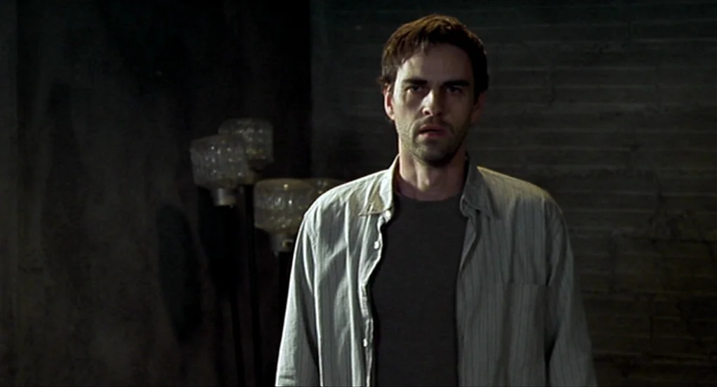 Tiresia (2003) movie still: medium — A dimly lit room with dark walls and a textured surface. A man in an open shirt stands before a three-headed…