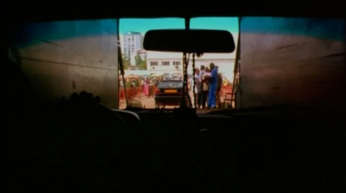 Code Unknown (2000) movie still: wide — View from inside a dark vehicle looking out through the front windshield at an outdoor area. A crowd of…
