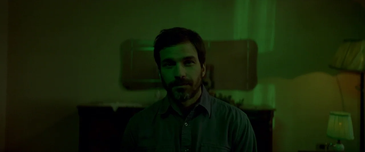 Ema (2019) movie still: medium — A man sits in a dimly lit room bathed in green light. He stares forward while the background remains obscured by…