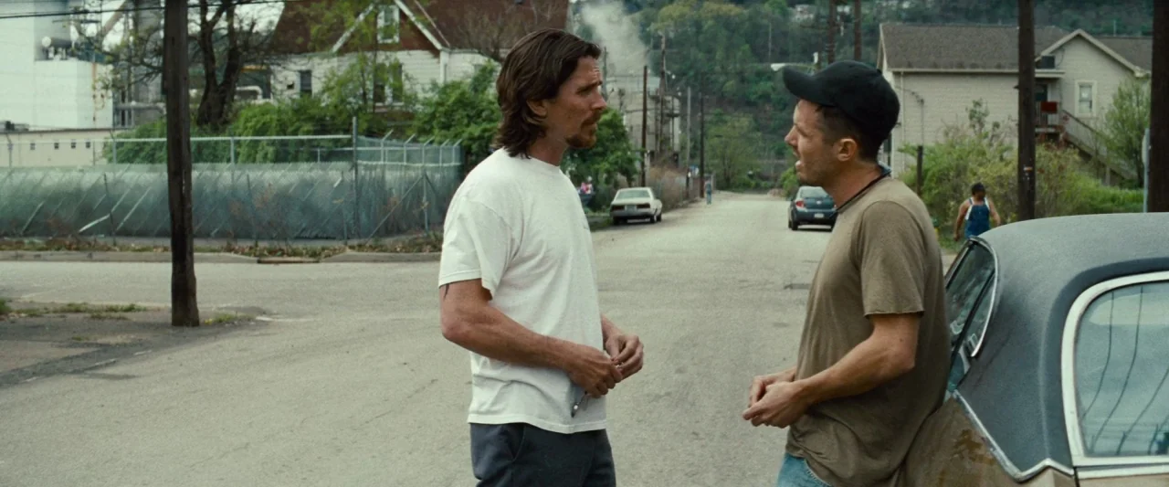 Out of the Furnace (2013) movie still: fullbody — A residential street intersection features houses and a metal chain-link fence. Two men converse…
