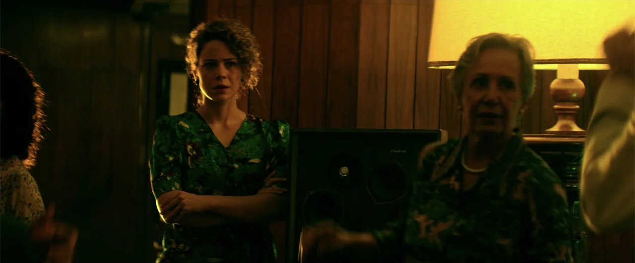 Bingo: The King of the Mornings (2017) movie still: medium — A dimly lit room with wood-paneled walls and a table lamp. Two women stand side-by-side…