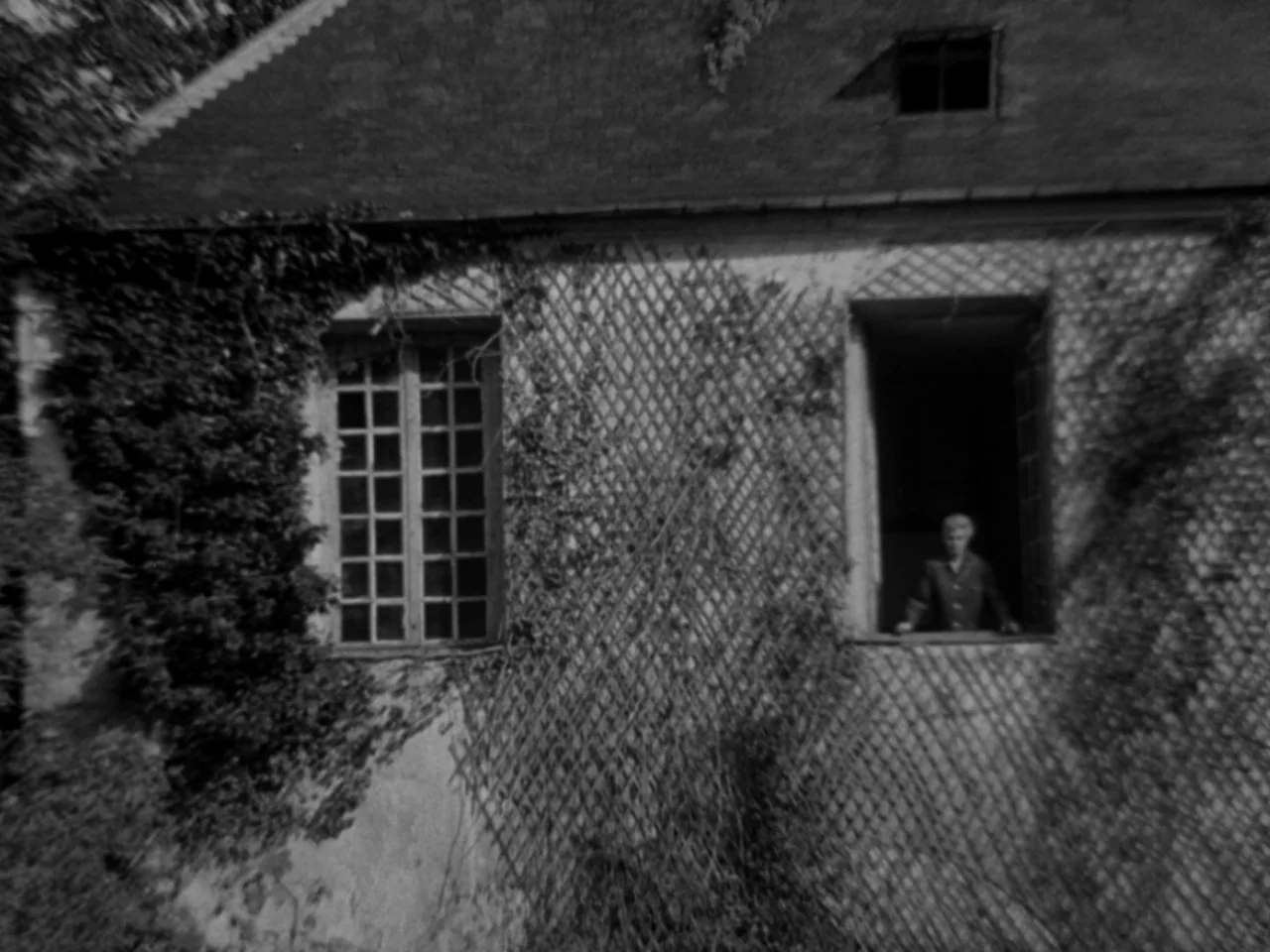 Diabolique (1955) movie still: wide — An exterior stone wall of a building is covered in a wooden lattice trellis. A young boy stands inside an open…