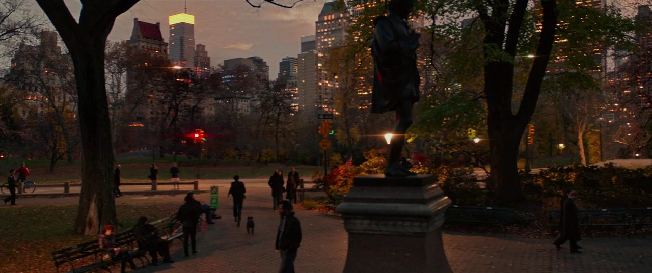 John Wick: Chapter 2 (2017) movie still: wide — A paved path winds through a park at dusk with city skyscrapers visible behind. Numerous pedestrians…