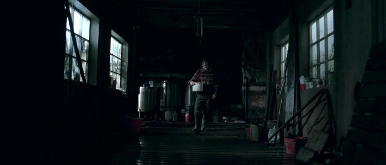 Isolation (2005) movie still: wide — A dark, industrial milking parlor with stainless steel equipment and scattered plastic buckets. A young man…