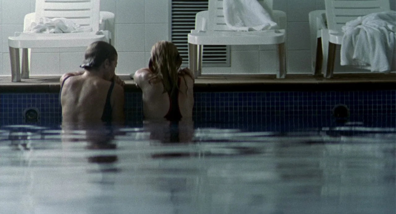 The Nameless (1999) movie still: medium — An indoor swimming pool with white plastic lounge chairs placed along the tiled edge. Two individuals in…