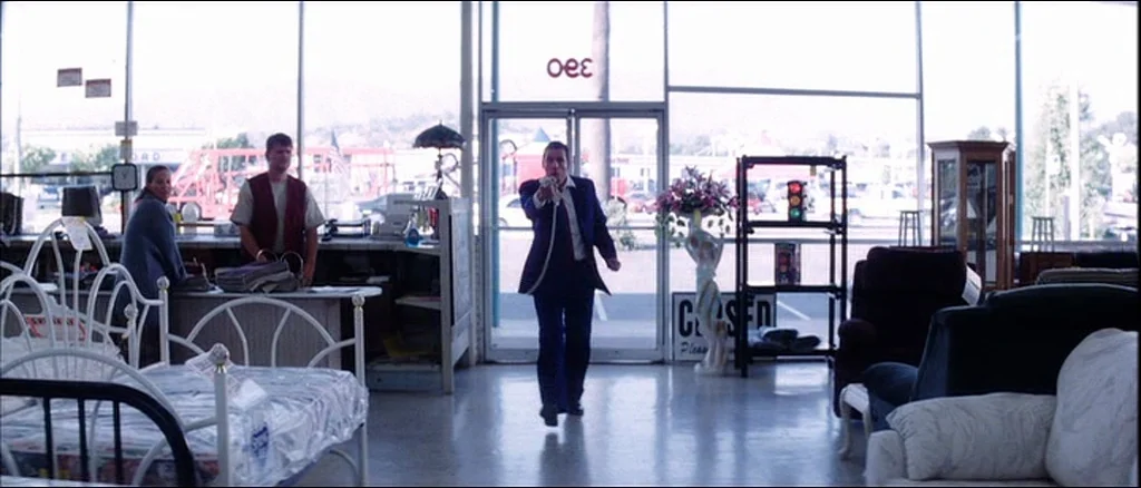 Punch Drunk Love (2002) movie still: fullbody — A furniture store interior features rows of mattresses and chairs visible under bright daylight. A…