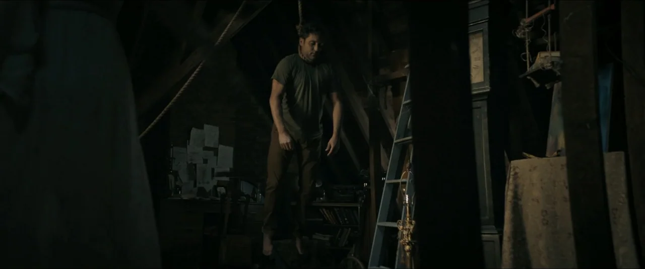 It Feeds (2025) movie still: fullbody — A cluttered, dark wooden attic contains a large grandfather clock and scattered papers. A man is suspended…