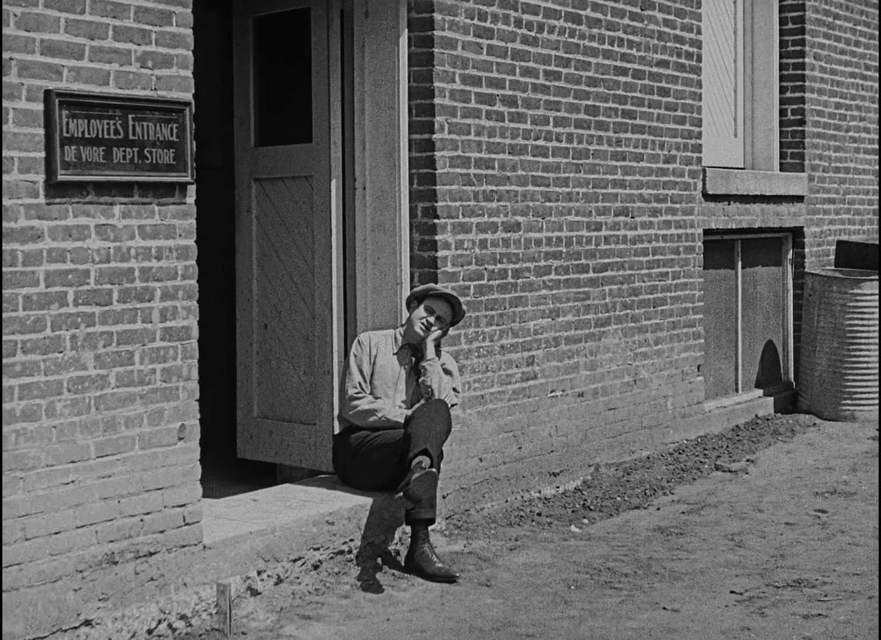 Safety Last! (1923) movie still: fullbody — A brick building exterior features a wooden door frame and a metal sign. A man sits on the concrete…