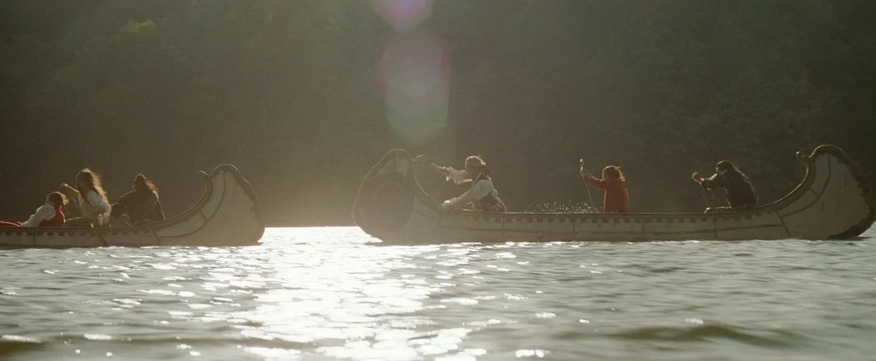 The Last of the Mohicans (1992) movie still: wide — Two birch bark canoes carry men across a sun-drenched river near a wooded bank. The subjects…
