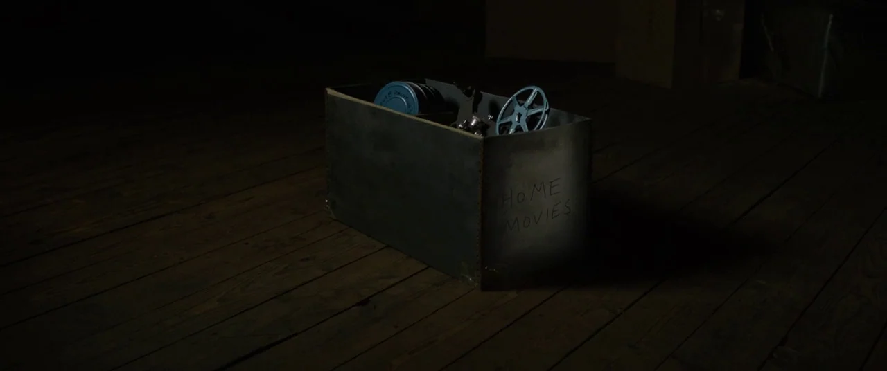 Sinister (2012) movie still: wide — A dark, wood-floored space contains a single cardboard box labelled home movies. The box contains several metal…