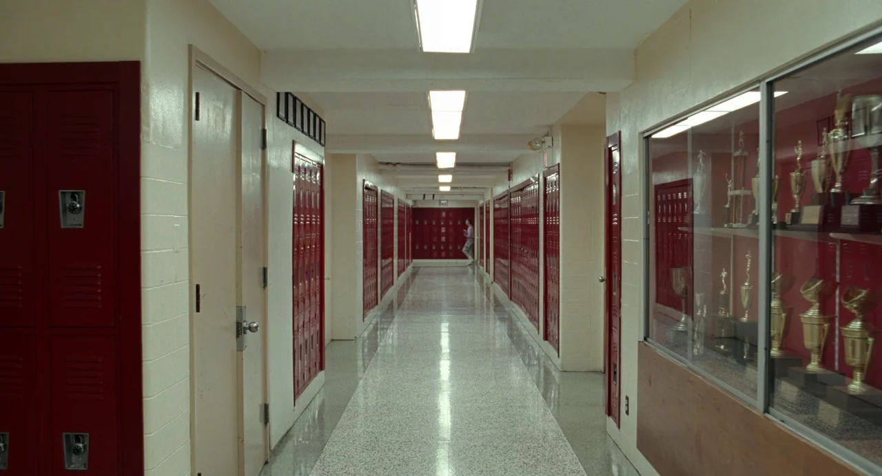Bones and All (2022) movie still: wide — A long school corridor features rows of red metal lockers and beige brick walls. A single, distant figure…