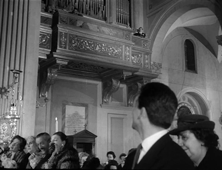 I Vitelloni (1953) movie still: medium — The interior of a stone church features an ornate carved wooden organ loft balcony. People in formal 1950s…