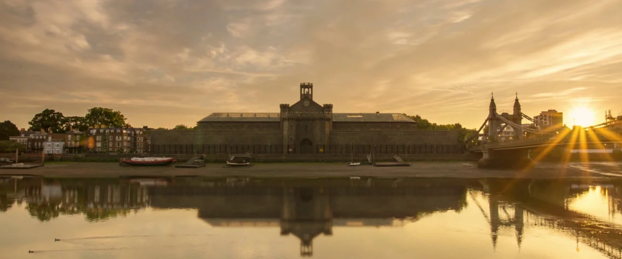 Paddington 2 (2017) movie still: establishing — A stone building with a central tower stands along a muddy riverbank at dusk. A bridge spans the…
