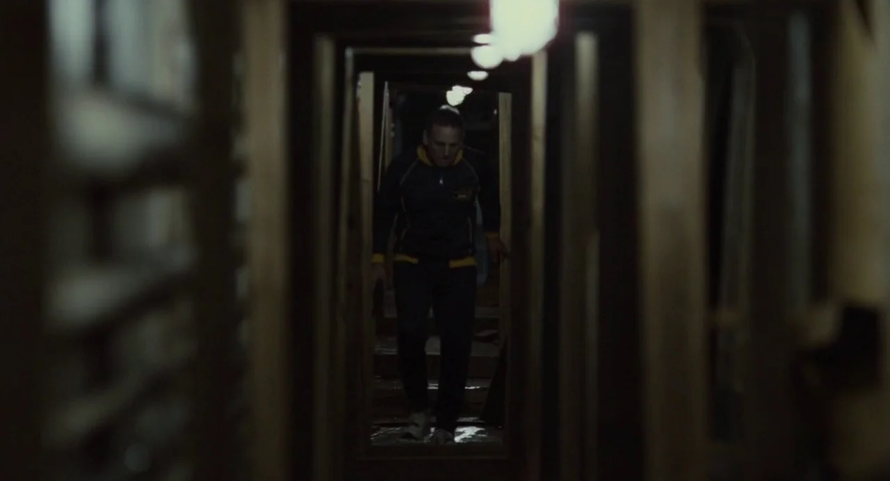 Foxcatcher (2014) movie still: medium — A long, dark, narrow corridor is defined by wooden vertical beams on both sides. A man in a dark tracksuit…