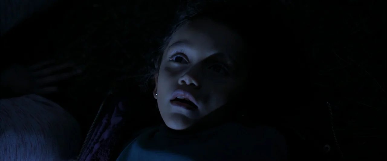 Evil Eye (2020) movie still: closeup — A dark outdoor space with grass visible in the shadows at night. A young child with curly hair looks upward…