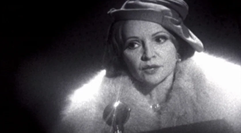 The Saddest Music in the World (2003) movie still: closeup — A woman stands in a darkened room with a single microphone in front of her. She wears a…