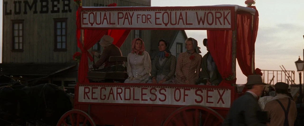 Tombstone (1993) movie still: wide — A group of women in period clothing sit on a red horse-drawn wagon. Large white banners on the cart display…