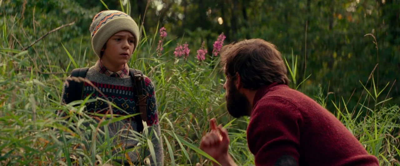 A Quiet Place (2018) movie still: medium — A man and a boy stand among tall grasses and wildflowers in a field. The man crouches slightly while…