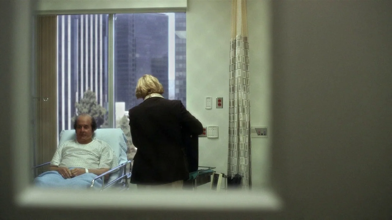 Behind the Candelabra (2013) movie still: medium — A hospital room features a window looking out onto city high-rise buildings. A man sits in a…