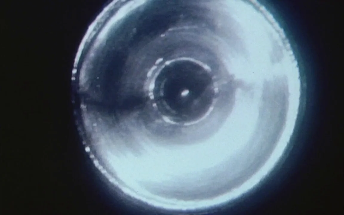 The Last of England (1987) movie still: closeup — A circular metallic aperture is centered against a dark, solid background. Light reflects off the…